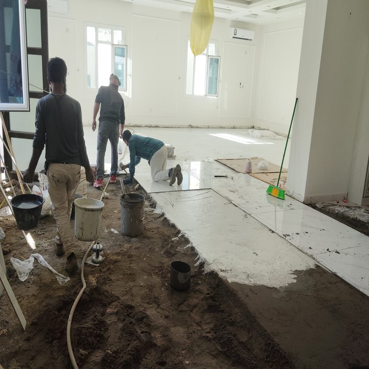 Workers are in the process of laying down flooring in an indoor construction site, preparing the ground and applying adhesive